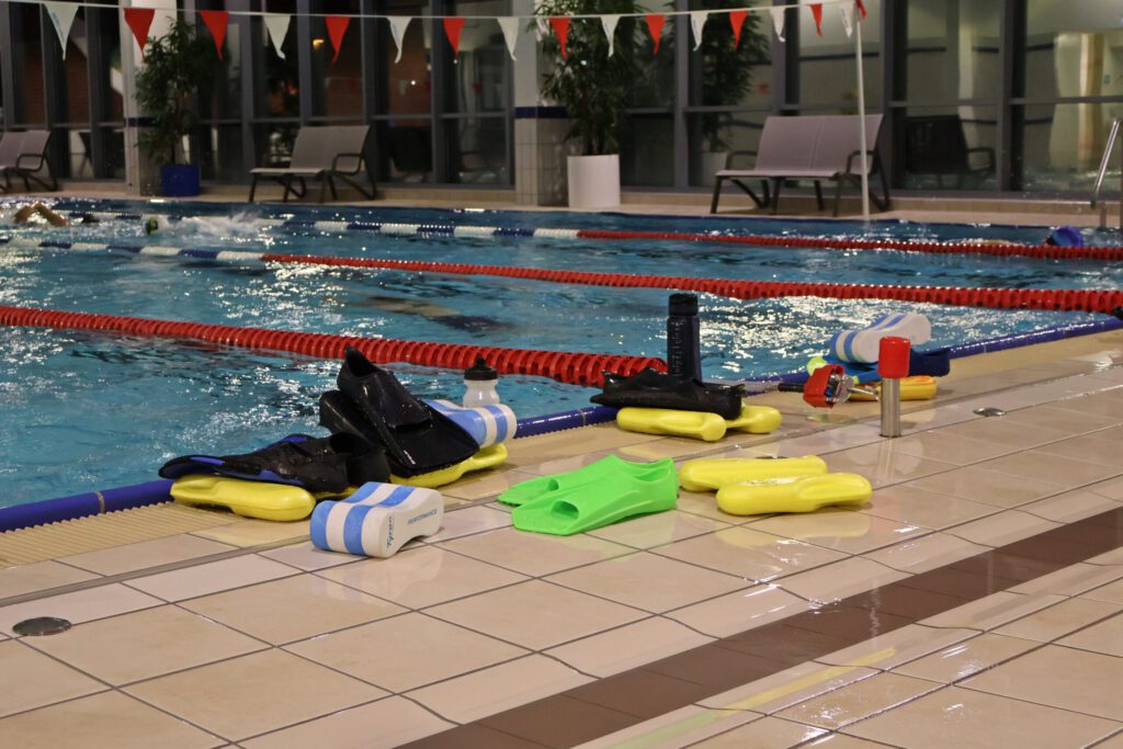 Mehrere Schwimmutensilien (Flossen, Bretter, Trinkflaschen) stehen am Beckenrand.