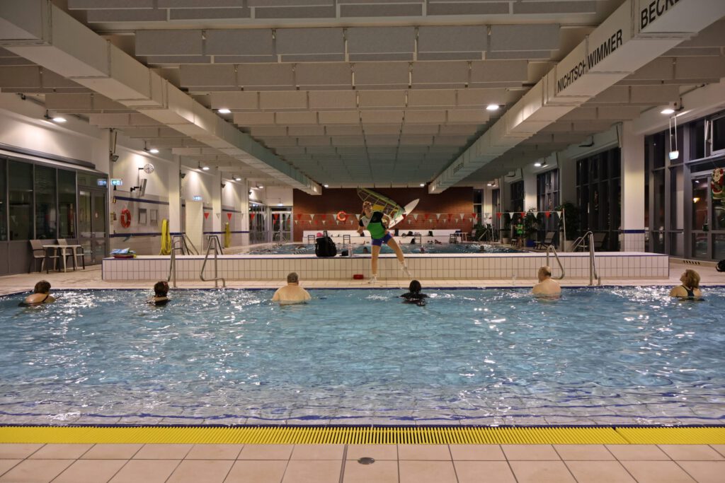 Aquafitnesstraining. 6 Personen im Wasser, eine Trainerin macht Übungen am Beckenrand vor